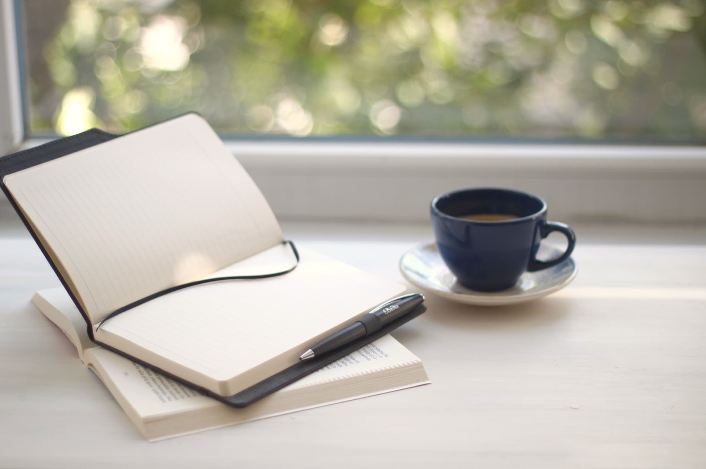 Cuaderno abierto sobre un libro cerrado, junto a una taza de café azul marino, frente a una ventana con luz suave.