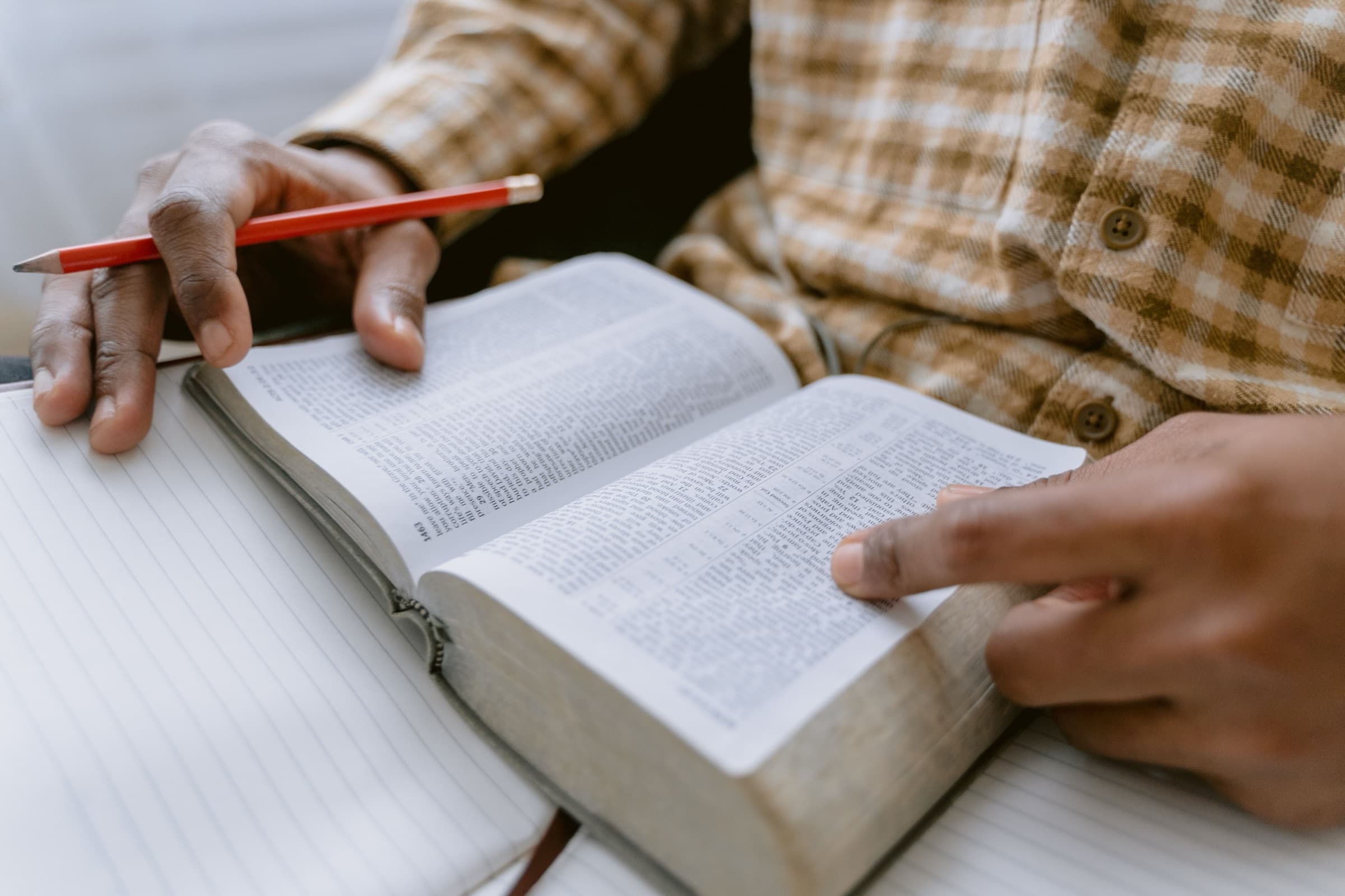 Manos señalando un versículo en una Biblia abierta, con un lápiz y un cuaderno sobre la mesa.